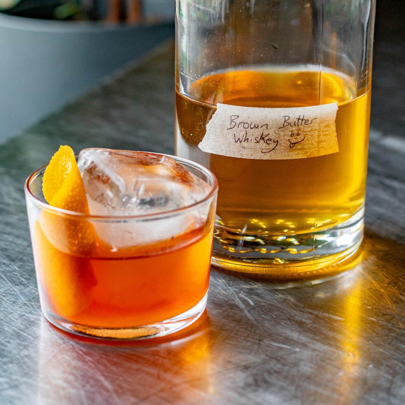 A tumbler with a big block of ice filled with a cocktail and a jug of brown butter whiskey behind